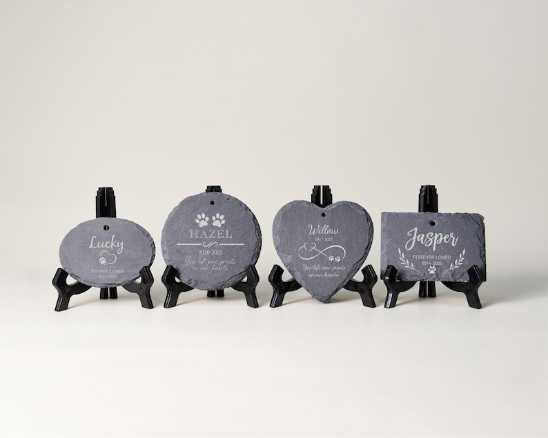 May include: Four slate pet memorial stones in oval, round, heart, and rectangular shapes, each on a black stand. The stones are engraved with pet names, dates, paw prints, and memorial text. The text is in a white color.
