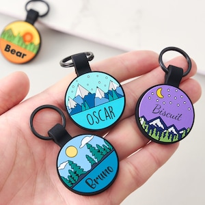 May include: A selection of round pet tags with black key rings. Each tag displays a different landscape design with a name: "Bear", "Oscar", "Bruno", and "Biscuit". The tags have a black border and a black strap.