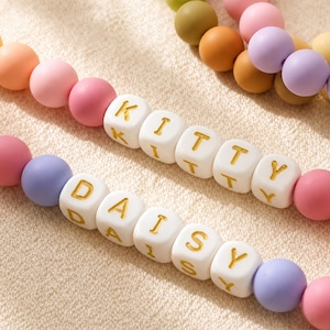 May include: Two strands of colorful matte beads featuring white square beads with gold lettering. One strand spells out the word KITTY and the other strand spells out the word DAISY. The beads are arranged in a repeating pattern of pastel colors.