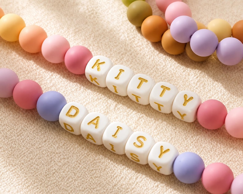 May include: Two strands of colorful matte beads featuring white square beads with gold lettering. One strand spells out the word KITTY and the other strand spells out the word DAISY. The beads are arranged in a repeating pattern of pastel colors.