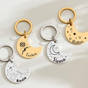May include: Four crescent moon-shaped keychains with gold and silver rings. Two are gold-colored with black designs and text, one with stars and the name "Button". The other gold keychain has a mountain design and the name "Tucker". The other two are silver-colored with floral and celestial designs and the names "Rosie" and "Sansa".