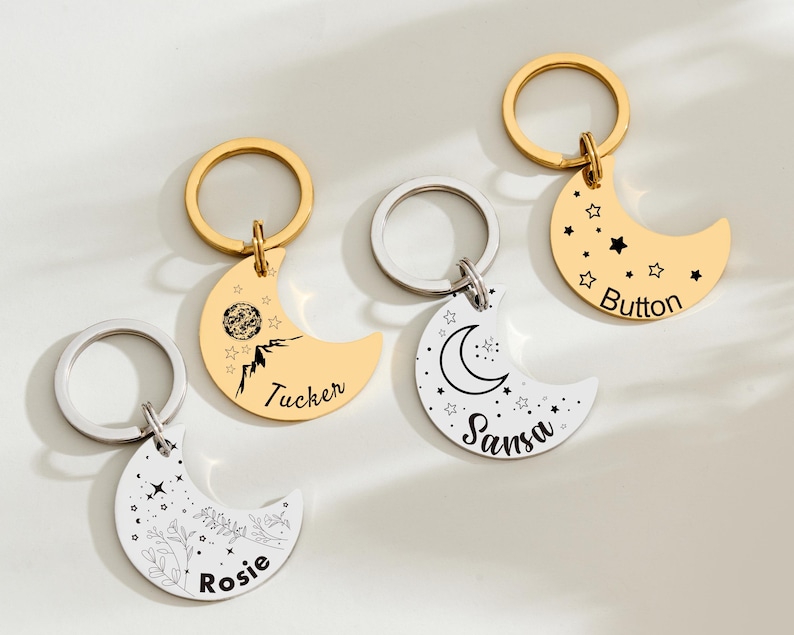 May include: Four crescent moon-shaped keychains with gold and silver rings. Two are gold-colored with black designs and text, one with stars and the name "Button". The other gold keychain has a mountain design and the name "Tucker". The other two are silver-colored with floral and celestial designs and the names "Rosie" and "Sansa".