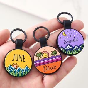 May include: Three circular keyrings with black loops. Each keyring displays a colourful landscape design with text. One keyring says "June" on a yellow background, another says "Dixie" on an orange background, and the last says "Scarlet" on a purple background.