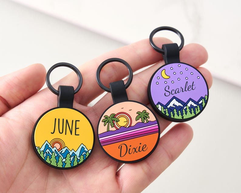 May include: Three circular keyrings with black loops. Each keyring displays a colourful landscape design with text. One keyring says "June" on a yellow background, another says "Dixie" on an orange background, and the last says "Scarlet" on a purple background.