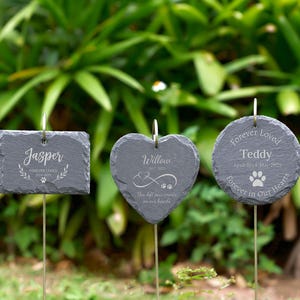 May include: Three dark gray slate garden markers with silver metal stems. One is rectangular, one heart-shaped, and one round. Each has engraved text: "Jasper", "Willow", and "Teddy", with dates and paw prints. The markers are in a garden setting.