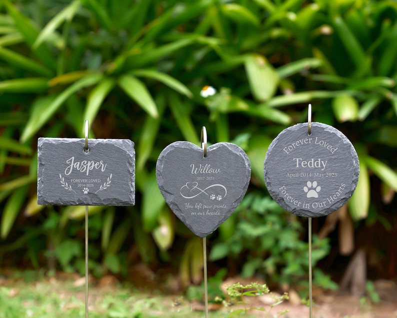 May include: Three dark gray slate garden markers with silver metal stems. One is rectangular, one heart-shaped, and one round. Each has engraved text: "Jasper", "Willow", and "Teddy", with dates and paw prints. The markers are in a garden setting.