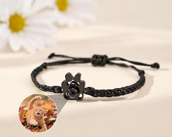 May include: Black braided bracelet featuring a paw-shaped charm with a small, round insert. The bracelet has an adjustable sliding knot closure. A small inset image shows a ginger cat in autumn leaves.