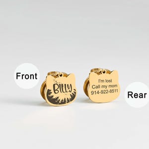 May include: Two gold-colored cat-shaped pet ID tags. One tag has the name "Billy" and a forest design. The other tag says "I'm lost Call my mom 914-922-8511". Both tags have a gold ring for attachment.