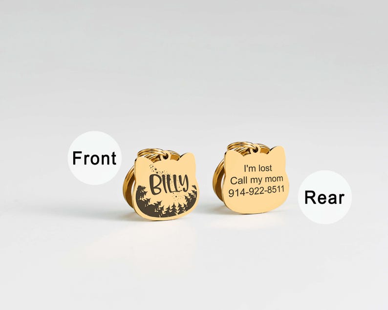 May include: Two gold-colored cat-shaped pet ID tags. One tag has the name "Billy" and a forest design. The other tag says "I'm lost Call my mom 914-922-8511". Both tags have a gold ring for attachment.