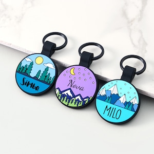 May include: Three circular keyrings with black loops. Each keyring has a different landscape design and a name. One features a sun, trees, and the name "Simba". Another has a moon, stars, mountains, and the name "Nova". The third has mountains and the name "Milo".