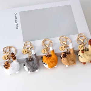 Peut inclure: Cinq porte-clés chats au crochet, blancs, gris, orange, marron et jaunes. Chaque chat a un porte-clés doré et une petite étiquette dorée avec un nom. Les chats sont dans une pose ludique, la queue en l'air.