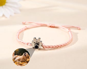 May include: A pink braided bracelet with a silver paw print charm. The charm has a small, round black center. A circular image of a golden retriever dog is next to the bracelet.