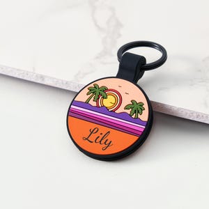 May include: A circular keyring with a black loop and holder. The keyring displays a sunset scene with palm trees, a sun, and the name "Lily" in black text. The background incorporates orange, purple, and pink hues.