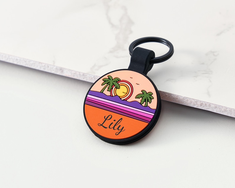 May include: A circular keyring with a black loop and holder. The keyring displays a sunset scene with palm trees, a sun, and the name "Lily" in black text. The background incorporates orange, purple, and pink hues.
