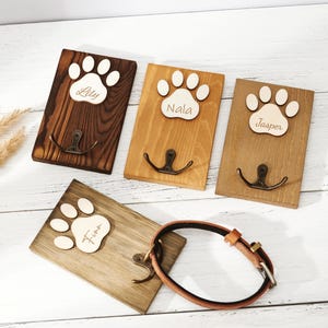 Puede incluir: Cuatro ganchos de pared rectangulares de madera para accesorios de mascotas, cada uno con una huella de pata de madera clara grabada con los nombres Lily, Nala, Jasper o Finn, y un gancho de metal oscuro fijado en el centro de la tabla.