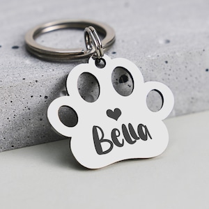 Könnte beinhalten: Ein silberfarbener Haustieranhänger in Form eines Pfotenabdrucks. Der Anhänger hat ausgeschnittene Kreise und ein Herz, mit dem Namen "Bella" in schwarzer Schrift. Ein silberner Ring ist befestigt.