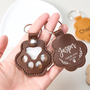May include: Brown leather paw-shaped keychains with silver keyrings. One keychain has a clear window, the other has the name "Jasper" and the text "Forever Loved 2014-2025". A third keychain is tan and has the name "Charlie".