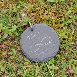 May include: A round, gray slate pet memorial marker with the name "Willow" and dates 2017-2025 engraved in a script font. The marker features an infinity symbol and paw prints, with the text "You left paw prints on our hearts."