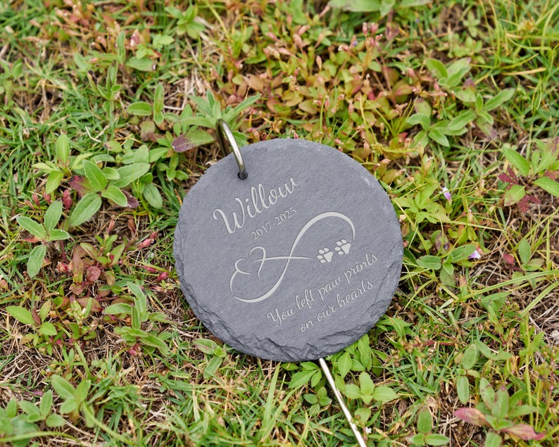 May include: A round, gray slate pet memorial marker with the name "Willow" and dates 2017-2025 engraved in a script font. The marker features an infinity symbol and paw prints, with the text "You left paw prints on our hearts."