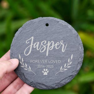 May include: A round, gray slate memorial stone with the name "Jasper" in elegant script. The text "FOREVER LOVED 2014-2025" and a paw print are also engraved. A small hole is at the top for hanging.