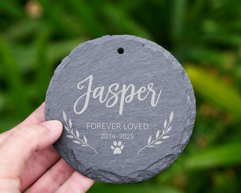 May include: A round, gray slate memorial stone with the name "Jasper" in elegant script. The text "FOREVER LOVED 2014-2025" and a paw print are also engraved. A small hole is at the top for hanging.