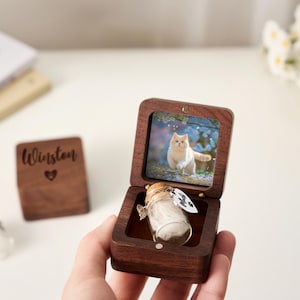May include: A wooden keepsake box with a photo of a cat inside. The box is open, revealing a small glass vial filled with white fluff. A small wooden square with the name "Winston" and a heart is also visible.