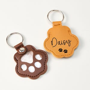 May include: Two paw-shaped keychains. One is brown with a clear center, the other is tan with the name "Daisy" and paw prints. Both have silver key rings and brown leather accents.