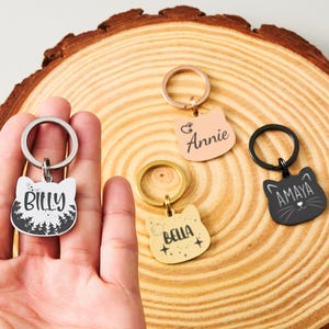 May include: Cat-shaped keychains in various colors, each with a name. One keychain is held in a hand. The keychains are silver, black, gold, and pink. The names on the keychains are Billy, Annie, Bella, and Amaya. The keychains are on a wooden surface.