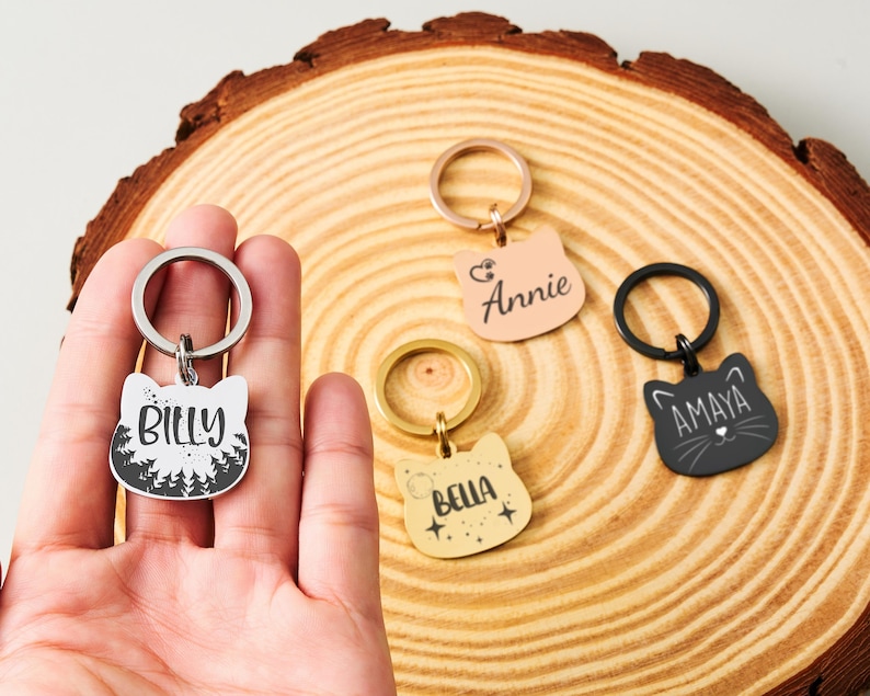 May include: Cat-shaped keychains in various colors, each with a name. One keychain is held in a hand. The keychains are silver, black, gold, and pink. The names on the keychains are Billy, Annie, Bella, and Amaya. The keychains are on a wooden surface.