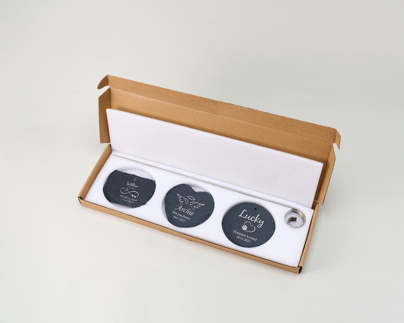 May include: A set of three dark gray slate pet memorial stones in a cardboard box. Each stone is engraved with a name and dates. The box is open, revealing the stones nestled in white foam.