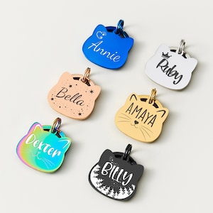 May include: A collection of cat-shaped pet ID tags in various colors, including blue, gold, and iridescent. Each tag features a different name, such as "Annie" and "Dexter", engraved in a stylish font. The tags have a metallic finish and a small ring for attachment.