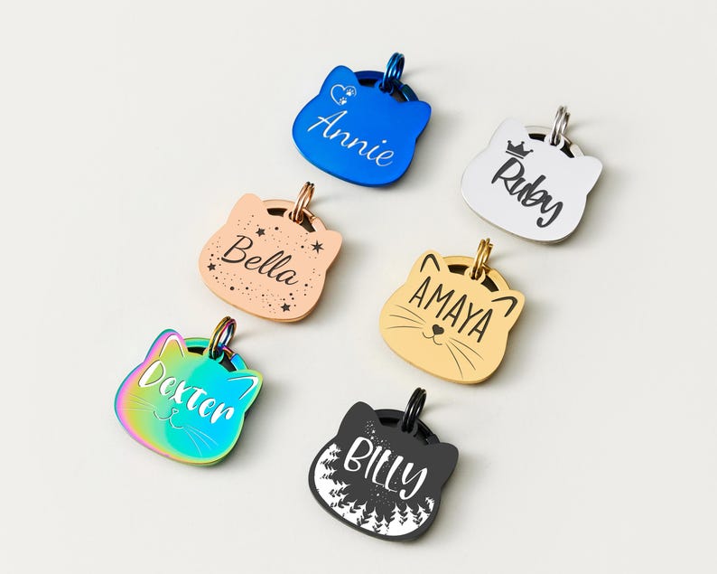 May include: A collection of cat-shaped pet ID tags in various colors, including blue, gold, and iridescent. Each tag features a different name, such as "Annie" and "Dexter", engraved in a stylish font. The tags have a metallic finish and a small ring for attachment.