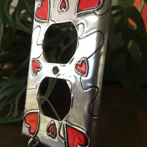 May include: A silver metal outlet cover with a decorative design of red hearts. The cover is designed to fit a standard double outlet.