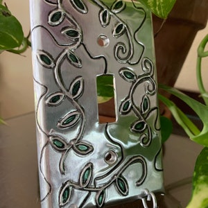 May include: A silver light switch cover with a decorative design of green leaves and vines. The cover has three small metal hooks on the bottom.