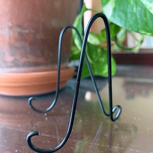 May include: A black metal display stand with a curved design. The stand has two curved supports and a base with curled ends. The stand is designed to hold a plate or other decorative item. The stand is on a reflective surface.