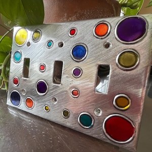 May include: A brushed metal light switch cover with three switch openings. The cover is decorated with colorful, circular accents in shades of yellow, teal, red, orange, blue, and purple. The design is modern and artistic.