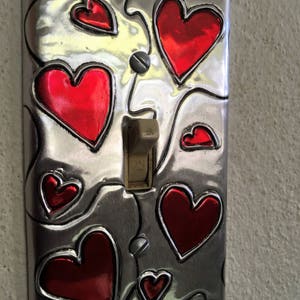 May include: A decorative light switch cover with a brushed metal finish. The cover features several red heart shapes outlined in black. The switch itself is a standard toggle style, and the word "ON" is visible.