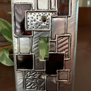 May include: A silver light switch cover with a geometric design featuring black and red glass squares. The cover is made of metal and has a textured surface.