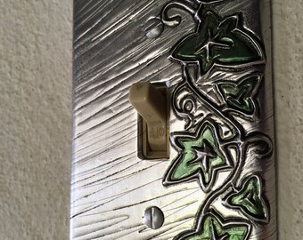 May include: A silver light switch cover with a decorative design of green ivy leaves. The switch is in the off position.