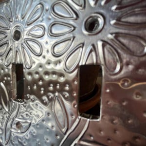 May include: A silver-colored, metal light switch cover with a floral design. The cover features two switch openings and is embossed with stylized flowers and a dotted texture. The design is symmetrical and detailed.