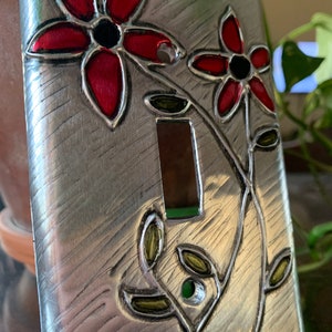 May include: A silver light switch cover with a red and green floral design. The flowers are stylized and have black centers. The switch cover has a brushed metal finish.