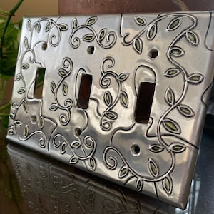 May include: A silver metal light switch cover with a decorative design of green leaves and vines.