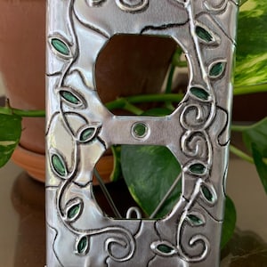 May include: A decorative silver metal outlet cover with a swirling vine and leaf design. The leaves are painted green. The cover has two openings for outlets and is displayed on a metal stand.