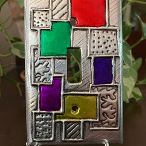 May include: A silver metal light switch cover with a geometric design. The cover features squares and rectangles in various colors, including red, green, purple, and yellow.