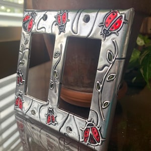 May include: A silver light switch cover with a double switch plate. The cover is decorated with a vine pattern and red ladybugs.