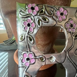 May include: A decorative silver-toned outlet cover featuring a floral design. Pink flowers with black centers and green leaves are painted on the cover. The cover has two outlet holes and is set against a blurred background.