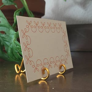 May include: A gold metal stand with a decorative design holds a blank card with a pink floral border. The card is on a brown wooden surface.