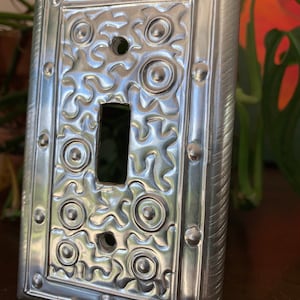 May include: A silver metal light switch cover with a decorative, embossed design of gears and circles. The cover has a brushed finish and a raised border.