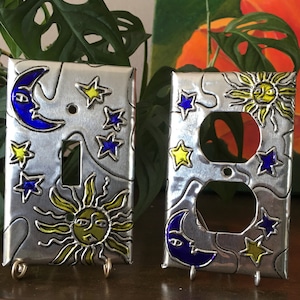 May include: Two decorative light switch covers with a celestial theme. One features a yellow sun with a face and blue stars on a silver background. The other features a blue crescent moon with a face and yellow stars on a silver background.