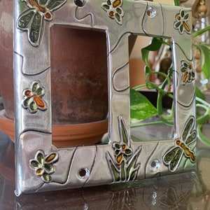 May include: A silver metal light switch cover with a dragonfly and floral design. The cover has a textured surface and is decorated with green, yellow, and orange paint.